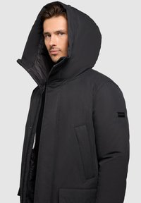 Muž se světlou pletí a světlýma očima, oblečený v černé prošívané bundě s velkou kapucí na hlavě, mírně se dívá doleva.