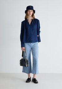 Blusa di lino blu navy con scollatura a V, jeans cropped blu chiaro, mocassini in pelle nera e un cappello a secchiello blu navy con logo. Borsa nera.
