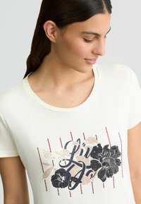 Crème T-shirt met korte mouwen met bloemmotieven in zwart en roze, verticale rode strepen, ronde halslijn en zachte stof.
