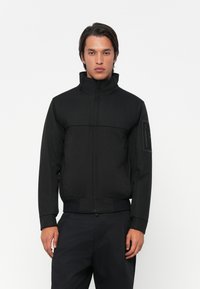 Black bomber jacket with high collar, zip closure, and ribbed cuffs. Features a side pocket with a textured patch on the left arm.