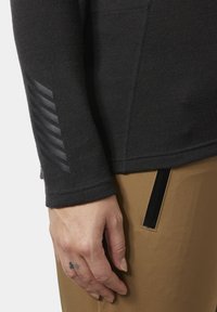 Detail ruky s malým tetováním, která má na sobě černý top s dlouhým rukávem s pruhy na rukávu a hnědé kalhoty s černým zipovým kapsou.