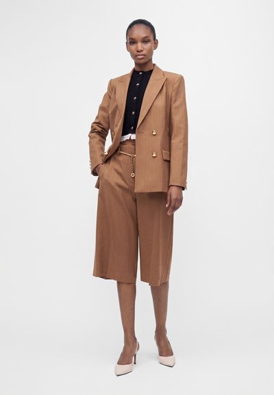 Femme portant un blazer beige à fines rayures avec des boutons dorés, un short assorti longueur genoux, un haut noir boutonné, une ceinture chaîne et des talons hauts beiges.