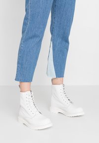 Stivaletti alla caviglia in pelle bianca con molteplici occhielli, punta arrotondata e suola spessa bianca. Indossati con jeans in denim azzurro chiaro con orlo spaccato.