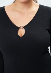 Schwarzes Strickoberteil mit V-Ausschnitt, das mit einem silbernen Ringakzent am Schlüsselbereich verziert ist. Glatte Textur und figurbetonte Silhouette.