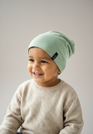 Smiling toddler wearing a light green beanie and beige sweater, looking slightly to the side against a plain, light background.