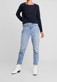 Pull marine, jean taille haute bleu clair, et bottines blanches à talon léger. Le pull a un col rond ; les jeans sont fuselés.