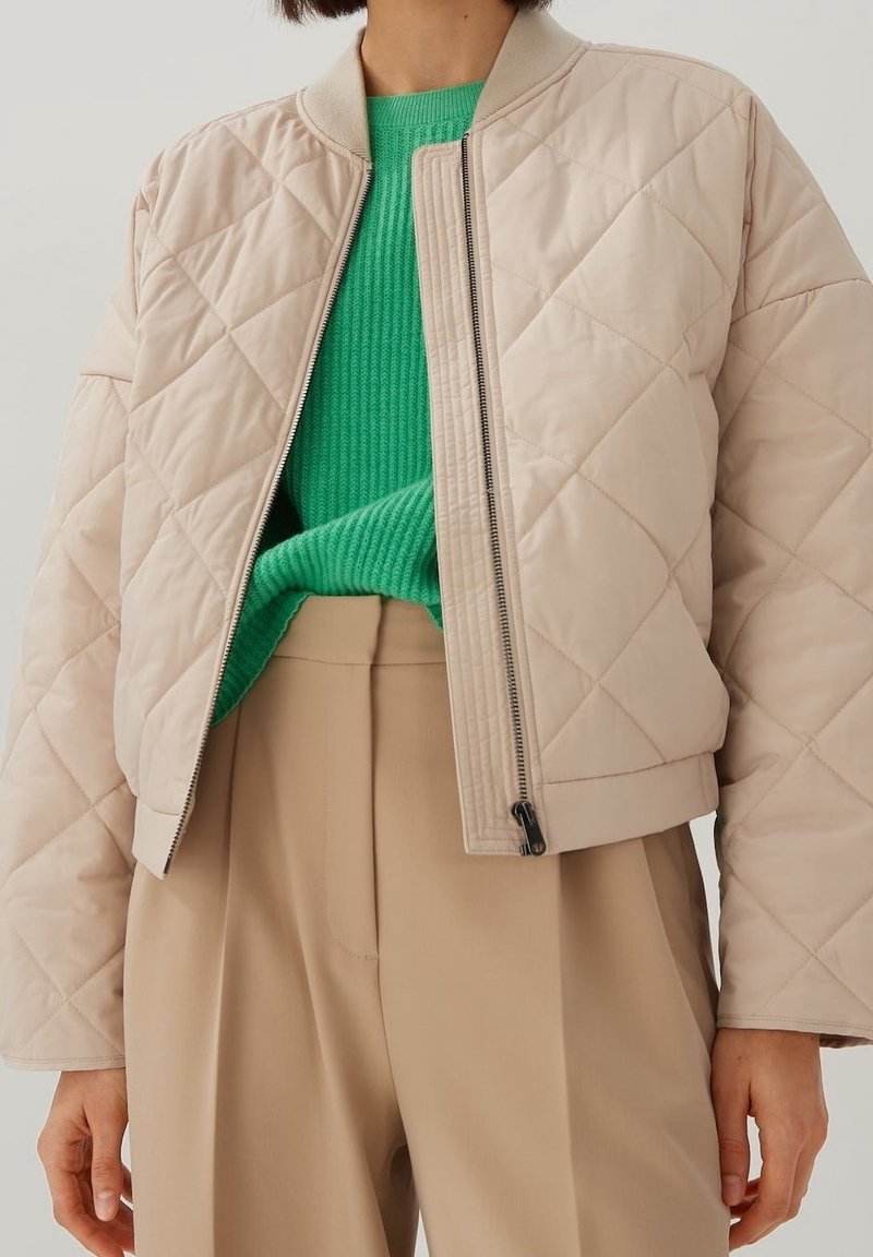 Personne portant une veste matelassée beige par-dessus un pull vert vif et un pantalon beige plissé taille haute sur un fond uni.