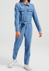 Salopette in denim con maniche lunghe, zip frontale, due tasche sul petto, vita con cintura e gambe affusolate. Colore azzurro chiaro con una texture liscia.