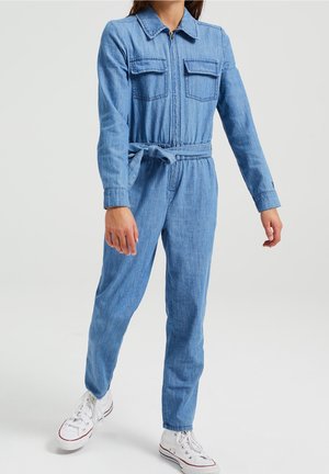Combinaison en denim à manches longues, fermeture éclair à l'avant, deux poches poitrine, taille ceinturée et jambes fuselées. Couleur bleu clair avec une texture lisse.