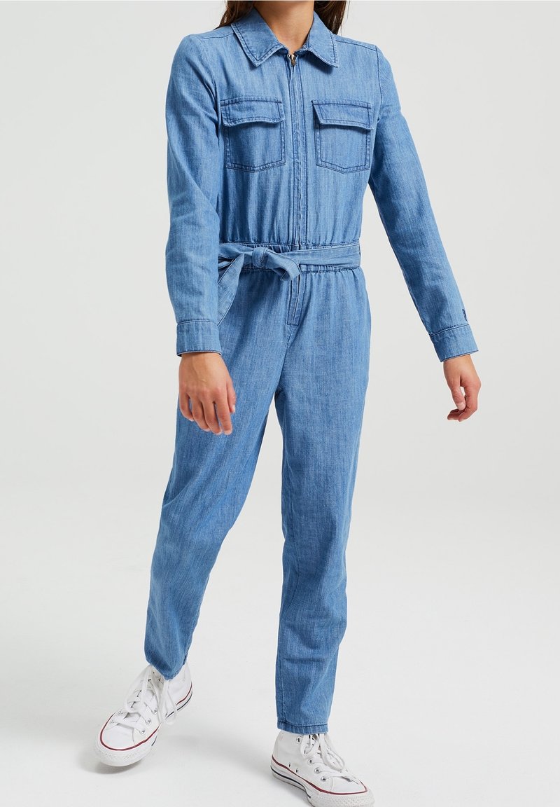 Salopette in denim con maniche lunghe, zip frontale, due tasche sul petto, vita con cintura e gambe affusolate. Colore azzurro chiaro con una texture liscia.