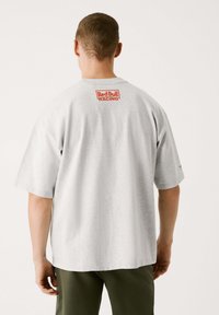 Graues Baumwoll-T-Shirt mit einem großen roten "Red Bull Racing"-Logo auf dem Rücken. Mit kurzen Ärmeln und lässigem Schnitt. Glatte Textur.