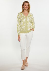 Hellgrüne Strickjacke mit weißem Blumenmuster, V-Ausschnitt, langen Ärmeln und Taschen, kombiniert mit weißen Hosen und hellen Schuhen.