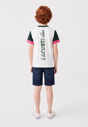 Enfant aux cheveux roux bouclés portant une chemise blanche avec des manches vertes et roses et un short en jean foncé, vu de dos sur un fond uni.