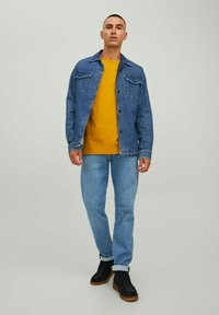 Chaqueta de mezclilla en azul medio con dos bolsillos en el pecho, llevada sobre una camiseta vibrante amarilla. Unos jeans azul claros y zapatos negros completan el atuendo.