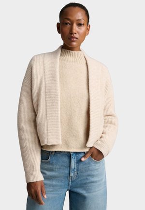 Frau trägt eine beige gerippte Strickjacke über einem beigen flauschigen Pullover und blauen Jeans, steht mit einer Hand in der Tasche vor einem schlichten Hintergrund.