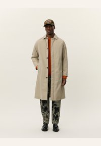 Palton bej cu croi drept, nasturi negri, asortat cu un pulover portocaliu și pantaloni închisi cu model, completat cu o pălărie verde.