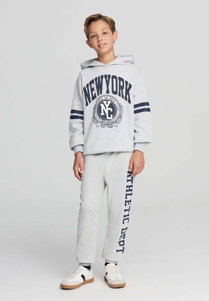 Felpa grigia con testo e strisce blu navy, abbinata a pantaloni della tuta coordinati con la scritta "ATHLETIC DEPT" stampata in verticale. Sneakers bianche con accenti scuri.