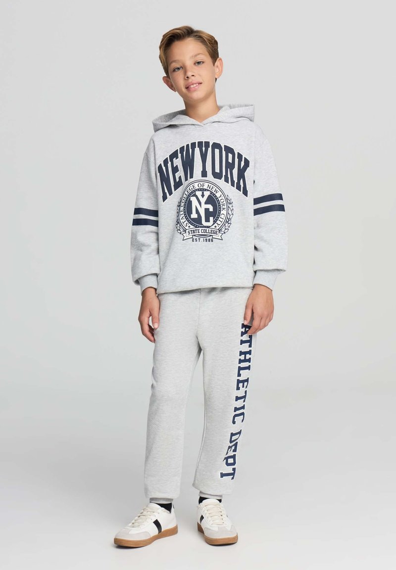 Felpa grigia con testo e strisce blu navy, abbinata a pantaloni della tuta coordinati con la scritta "ATHLETIC DEPT" stampata in verticale. Sneakers bianche con accenti scuri.