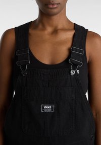 Zwarte denim overalls met verstelbare schouderbanden, een voorzak en een Vans-logo label op de borst. Glad textuur en minimalistisch ontwerp.