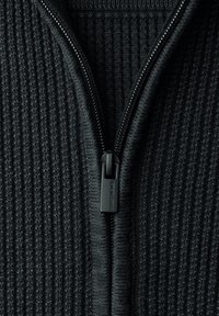 Tissu en maille côtelée noire avec une fermeture éclair noire partiellement zippée, dotée d'un tirette rectangulaire, sur un vêtement.