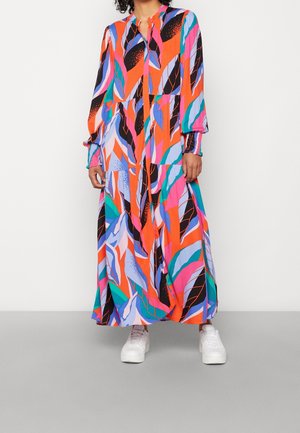 Kobieta ubrana w długą, kolorową sukienkę w abstrakcyjne wzory w odcieniach pomarańczy, różu, niebieskiego, fioletu i czerni, zestawioną z białymi trampkami.