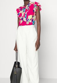 Květinová blůza v jasně růžové barvě s volánovými rukávy, spárovaná s bílými stylovými kalhotami. Černá kabelka se zlatými akcenty dotváří celek.