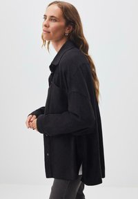 Camicia nera con bottoni, dal taglio rilassato, caratterizzata da maniche lunghe, colletto e una tasca frontale. Realizzata in un tessuto morbido, con una texture minima.