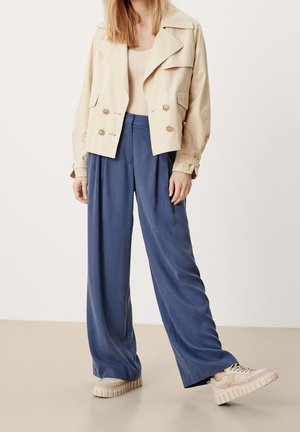 Personne portant une veste cropped beige, un haut en tricot crème, un pantalon bleu à jambes larges et des baskets à plateforme beiges se tenant sur un sol clair.