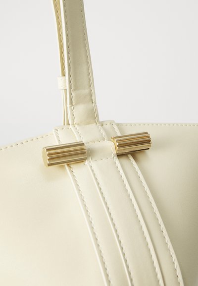 Kremkrāsas ādas rokassoma ar teksturētu virsmu, kurai ir centrālā sprādze ar zelta akcentiem un izšūtu apdari uz rokturīša.