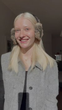 Šedý vlněný kabát, černý top, dlouhé blond vlasy, kožíškem lemované chrániče uší, usměvavý výraz, přirozené osvětlení zvýrazňující rysy.