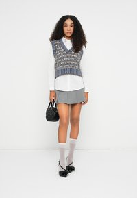 Falda corta gris y camisa blanca con cuello, superpuesta con un chaleco de punto azul con estampado. Los accesorios incluyen un pequeño bolso negro y medias altas blancas.