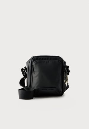 Zwarte compacte crossbodytas met voorvak met rits, verstelbare riem en gestructureerde stof tegen een effen achtergrond.