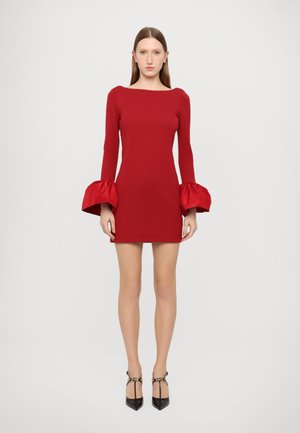 Vestido mini ajustado rojo con mangas largas que presentan puños voluminosos y con volantes. Textura suave con un escote cuadrado. Combinado con zapatos de tacón negros.
