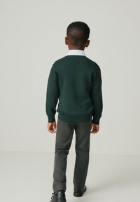 Dunkelgrüner Pullover mit rundem Ausschnitt, kombiniert mit grauer Hose. Das Outfit wird durch einen weißen Kragen und schwarze Schuhe ergänzt.