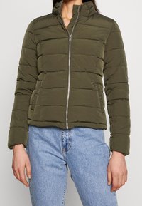 Olivengrøn dynejakke med høj krave, lynlåslukning og vandret quiltet mønster. Har to sidelommer og en kort pasform.