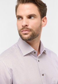 Camicia bianca con una fantasia a pois viola; dettagli notevoli includono un colletto classico e bottoni marroni. La texture appare liscia e tessuta.