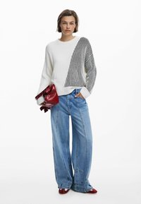 Witte geribbelde trui met een zwart gestreept diagonaal patroon, gecombineerd met lichtblauwe wijd uitlopende jeans en een kleine rode handtas.