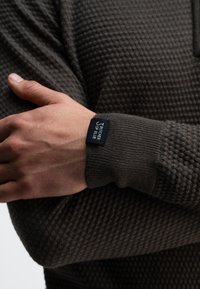 Wafeltextuur, donkerbruin sweater met geribde manchetten. Voorzien van een zwart label met witte tekst. De arm is gekruist en toont het kledingstuk.