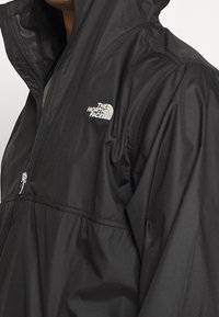 Zwarte windbreaker jas met halve rits, capuchon en wit "The North Face" logo op de linkerborst, gedragen door een persoon.