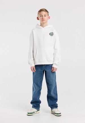 Adolescent debout, portant un sweat à capuche blanc avec un logo vert, un jean bleu et des baskets blanches avec des accents verts sur un fond clair uni.
