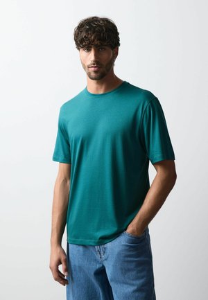 Mand med krøllet hår iført en turkis kortærmet skjorte og blå jeans står med den ene hånd i lommen mod en ensfarvet hvid baggrund.