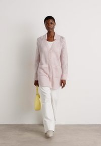 Lichtroze gebreide cardigan met een gestructureerd, open geweven design, twee voork 기반kante zakken en een kraag. Gecombineerd met witte brede broekspijpen en een gele tas.