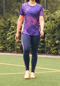 Camiseta deportiva morada con acentos en rosa, combinada con leggins azul marino que tienen rayas laterales rosas. Unos zapatos deportivos amarillos brillantes completan el conjunto.