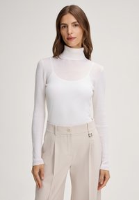 Weißes, geripptes Turtleneck-Oberteil mit langen Ärmeln und taillierter Passform, kombiniert mit hellbeigen, hochgeschnittenen Hosen mit Knopfdetail.