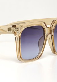 Ochelari de soare transparenti de culoare maro deschis, cu lentile pătrate, cu un gradient de nuanțe de violet închis la violet deschis. Rame groase cu finisaj strălucitor și articulații metalice.