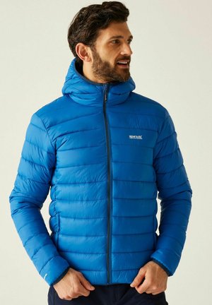 Hombre con chaqueta acolchada azul brillante con capucha, cerrada con cremallera, con las manos cerca de la cintura, mirando hacia la derecha contra un fondo claro y liso.