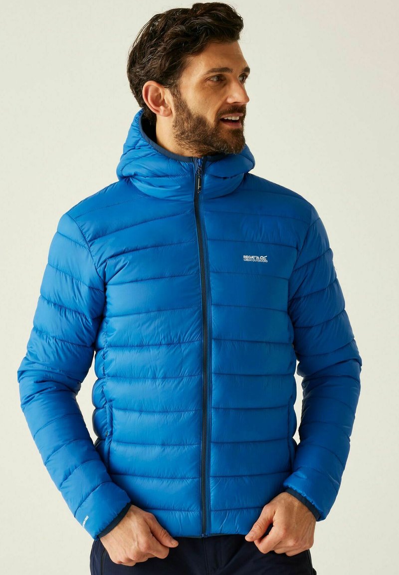 Hombre con chaqueta acolchada azul brillante con capucha, cerrada con cremallera, con las manos cerca de la cintura, mirando hacia la derecha contra un fondo claro y liso.