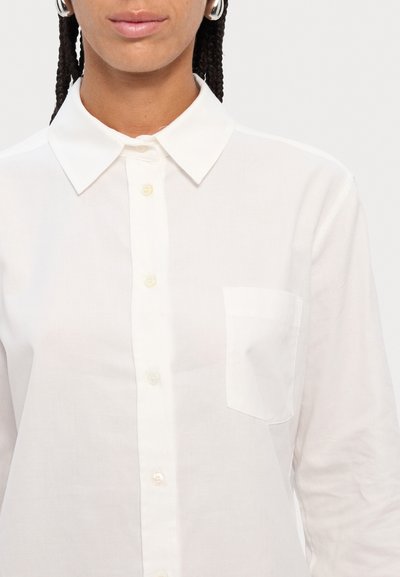 White long-sleeve shirt with a pointed collar, button placket, single chest pocket, and textured fabric. Buttons are light cream.