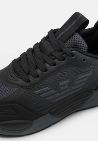 Svart idrottssko med nätpaneler, texturerat läder och tonade detaljer. Har runda snören och en vadderad krage för stöd.