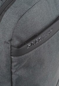 Mochila de tela gris con un destacado logo negro de Skechers, superficie texturada y un compartimento con cremallera. Diseño compacto y moderno.